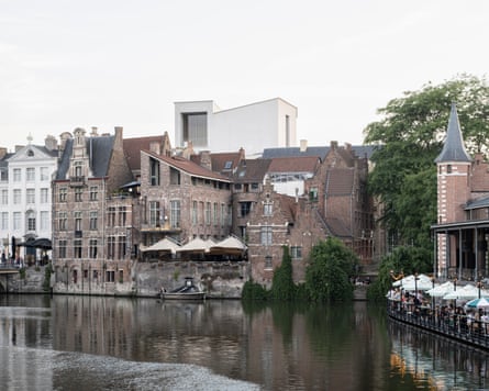 Historic buildings in Ghent line a canal, with diners seated under umbrellas beside the water, with the boxy white museum emerging above the rooftops