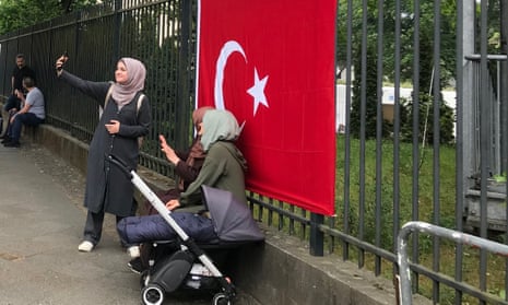 Three women with a buggy in front of a Turkey flag hung on a railing taking selfies.