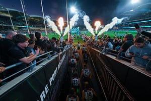 Australian rules football players walk up along a crowd-lined tunnel to the pitch as fireworks go off