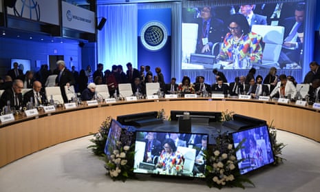 Delegates from various countries sit at a circular desk as one of the speakers is shown on a large screen