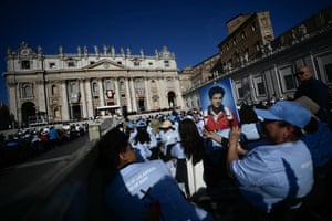 A group of people sitting in an ornate square, one of them holding up an image of Carlo Acutis