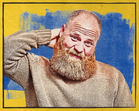 A bald-headed man with a beard wearing a taupe-coloured jumper, with his hand behind his neck