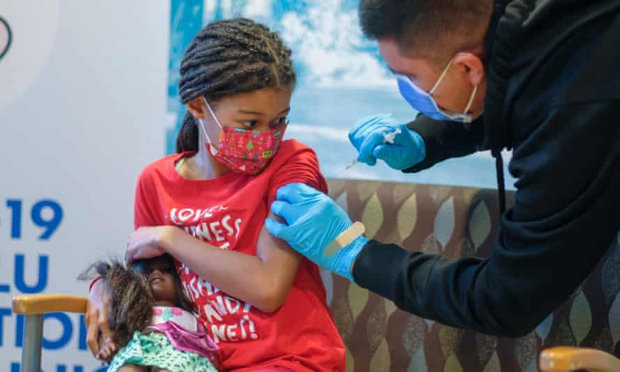 A 7-year-old African-American girl  wearing a read t-shirt and mask, and holding an African-American doll, receives a Covid-19 vaccine from a gloved, masked health care worker.