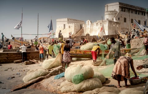 A bustling harbour scene with people tending to nets and long traditional fishing boats in front of a colonial-era castle.