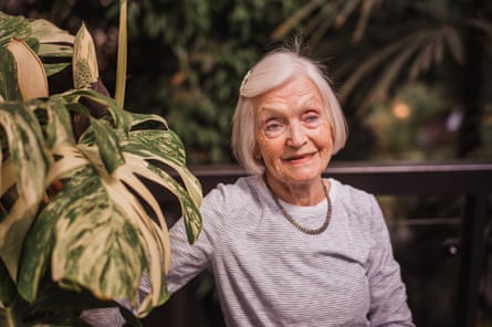 Stella, 88, stands next to a giant monstera. She has white hair in a bob fastened back to one side and wears a grey top with narrow stripes.
