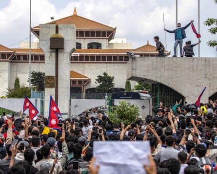 A crowd gathered in front of the building, some with flags and signs. Some people stand on a wall in front of the building