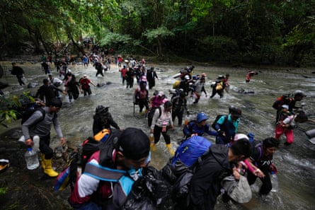 A large crowd of people carrying backpacks and water bottles wades across a wide river in the jungle.