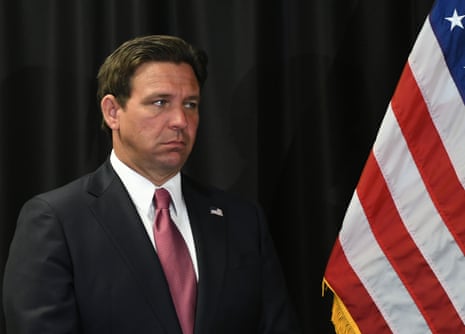 White man wearing black suit and red tie next to US flag