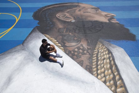 The mural artist Gustavo Zermeno Jr sits on a basketball court mural he dedicated to Nipsey Hussle in Los Angeles.