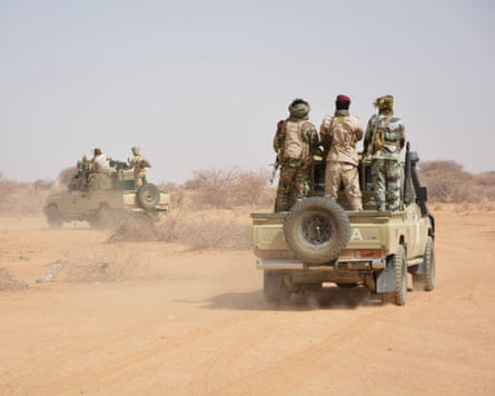 Men in uniform standng up in the back of military pickups driving through a desert landscape.