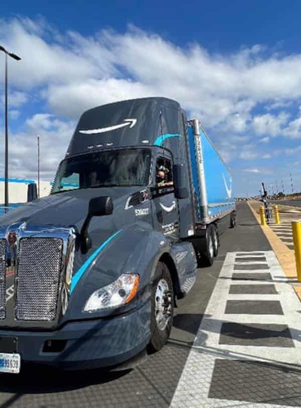 A gray and blue semitrailer truck with the Amazon logo approaches the camera, with a warehouse in the background.