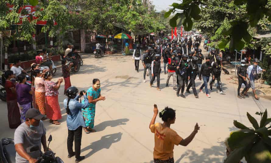 A group of black clad protesters carrying banners walk along a dusty street, cheered by a small group of residents at the side of the road.