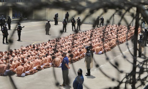 Prisoners sat on the cement floor of a courtyard at a prison. Their upper bodies are bare, their heads shaved, and they wear white shorts. They're surrounded by police officers with assault riffles. Some have '18' tattooed on their backs, signifying they are members of a gang.