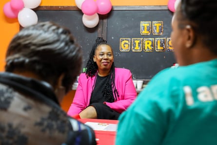 A woman in a pink jacket sits in front of a blackboard talking to two other women in the foreground