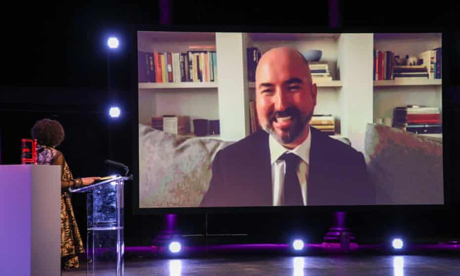 Douglas Stuart seen on a large illuminated screen on a dark stage, talking to a compere who is standing at a lectern.