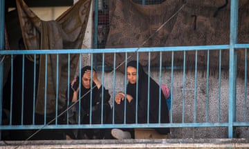 Two women crouch on balcony of damaged building