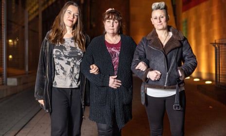 Three women standing in a street, linking arms. They are all wearing dark-coloured jackets, T-shirts and trainers. One has long brown hair, another has shorter brown hair and another has short blond hair combed upwards.