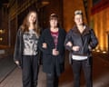 Three women standing in a street, linking arms. They are all wearing dark-coloured jackets and T-shirts and trainers. One has long brown hair, another has shorter brown hair and another has short blond hair combed upwards.