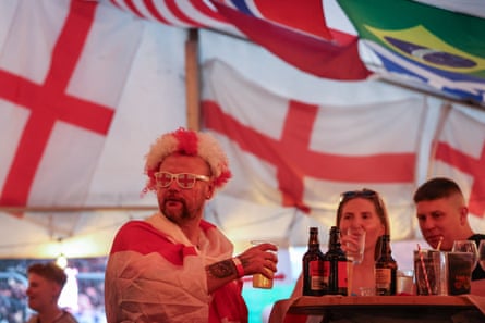 A fan wearing a wig and glasses in England colours and draped in the St George’s flag holds a pint alongside a table of drinks with two other fans