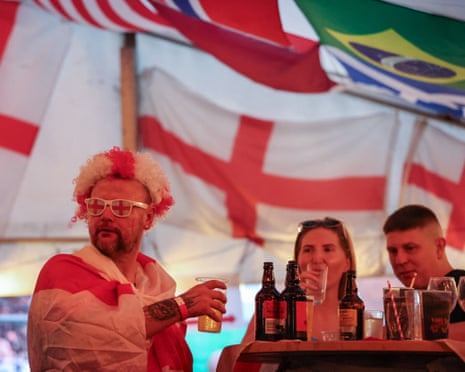 A man in a red and white wig and glasses holds a drink at a pub table with beer bottles