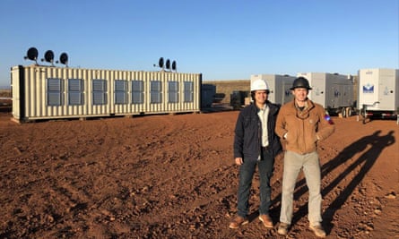 Two men stand in a dirt field next to a shipping container-like structure that is their bitcoin mining data center.