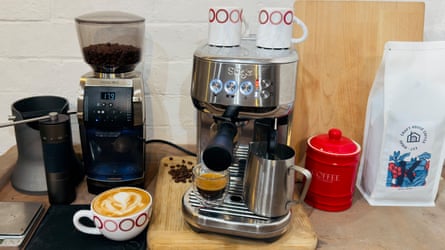 A sideboard with a silver coffee machine, black coffee grinder, a cup of coffee and a bag of coffee beans