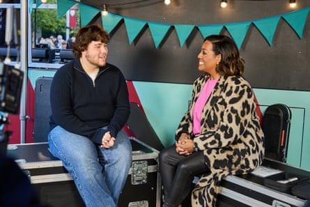 A young man with curly dark hair and beard talking to a woman in a cow-print coat who is smiling broadly - with bunting behind them