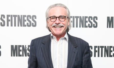 Middle-aged white man with combed-back white hair, bushy white moustache, and black round glasses, in suit jacket and shirt with no tie, smiles broadly in glaring lights in front of background that says “Men’s Fitness”.