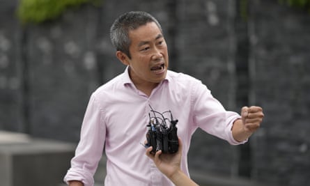 An animated middle-aged east Asian man gesticulates as he speaks to people off-camera. A hand holds microphones in front of him as he speaks. 