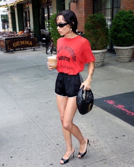 Zoë Kravitz in sunglasses, carrying iced coffee and an YSL handbag and wearing a red Black History Month T-shirt with black shorts and mules