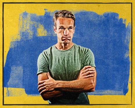A man in a green t-shirt stands with arms crossed and an uncertain expression against a blue and yellow background