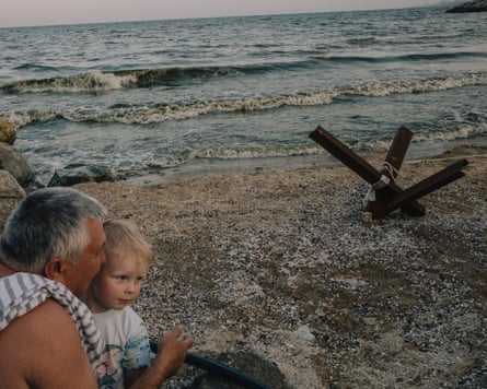 A man and child sit on a beach with a metal structure on the shore nearby.