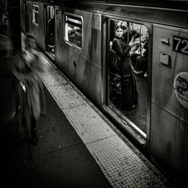 A black and white image of a subway train, with a woman at a carriage doorway looking out while another woman, blurred, walks the platform
