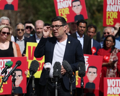 Andy Burnham speaks into a row of microphone as a crowd hold placards saying 'Vote Andy for us'