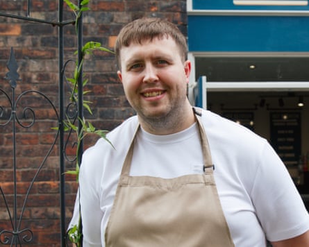 Kieran stands outside his cafe in a white t-shirt and chef’s apron