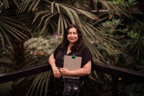 Lydia Goldberg stands in front of tall green palm-like trees. She is in her mid-50s and has long dark hair; she wears a black top and carries a bag with lots of pockets plus a clipboard.