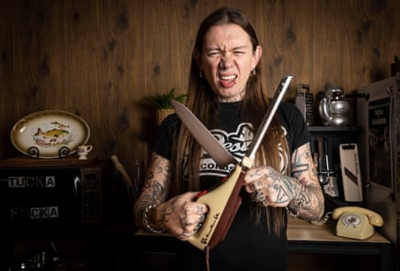 A man with long hair and tattooed arms and wearing a black T-shirt standing in a kitchen, holding a pair of crossed knives in front of 