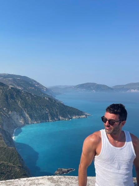 He poses against a wall, wearing a white tank top and dark sunglasses, against a beautiful coastal backdrop