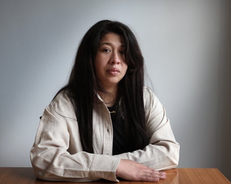 A woman with long dark hair sits at a wooden table, looking at the camera