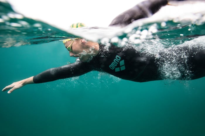 A split photo above and below the water of Kai Edwards swimming freestyle in the ocean. He is wearing a black full-body swimsuit and a yellow and green cap