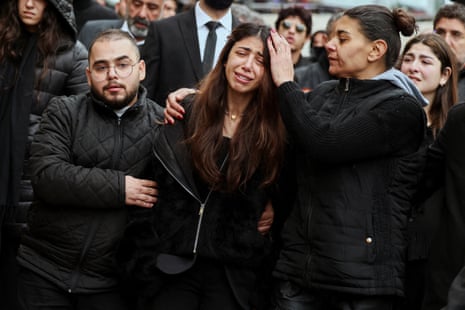 काले कपड़े पहने एक पुरुष और एक महिला काले कपड़े पहने एक रोती हुई महिला को संभालते हैं।