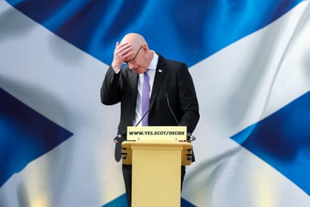 John Swinney stands at a lectern in front of a scottish flag with his head in his hand