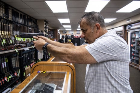 A man stands at a counter in a gun shop holding a firearm as if ready to shoot.