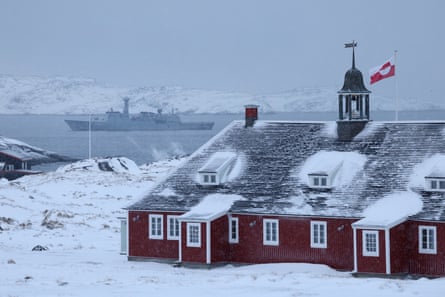 The Greenlandic flag flies over a building as the grey navy frigate is in the sea behind. Snow lies on the ground and on the building's roof