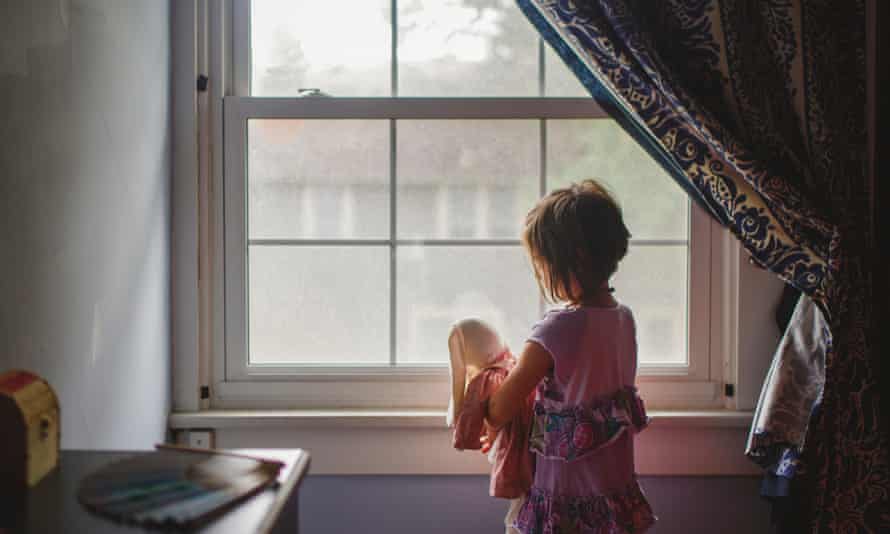 A small child stands by bedroom window tenderly holding stuffed bunny