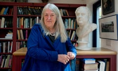 A smiling Mary Beard in her library at home in Cambridge, shelves full of books behind her, standing beside a bust of the Roman emperor Vitellius.