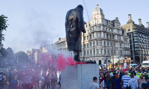 Red and white smoke billow from flares on the plinth of a statue of Winston Churchill surrounded by people.