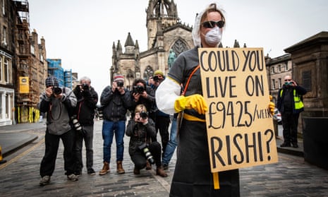 A young woman wearing a mask and gloves holds up a sign saying ‘Could you live on £94.25/week Rishi?’