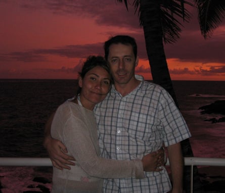 The couple stand with their arms around one another, with a palm tree behind them and the setting sun making the sky glow a dark orange