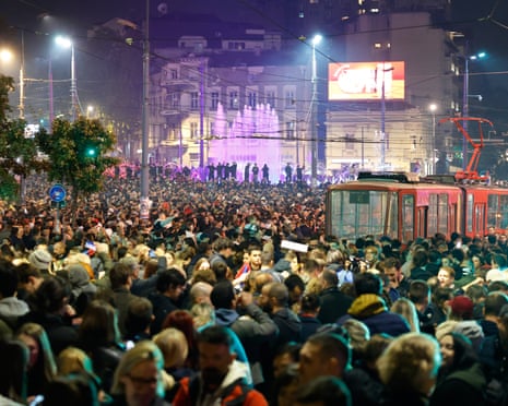 Crowds celebrate at night near a lit fountain in Belgrade, with a tram parked nearby
